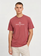 A man wearing a dusty rose short-sleeve t-shirt with a white gothic-style cross and 'jesus saves' text on the chest, paired with light beige pants, against a light gray studio background.