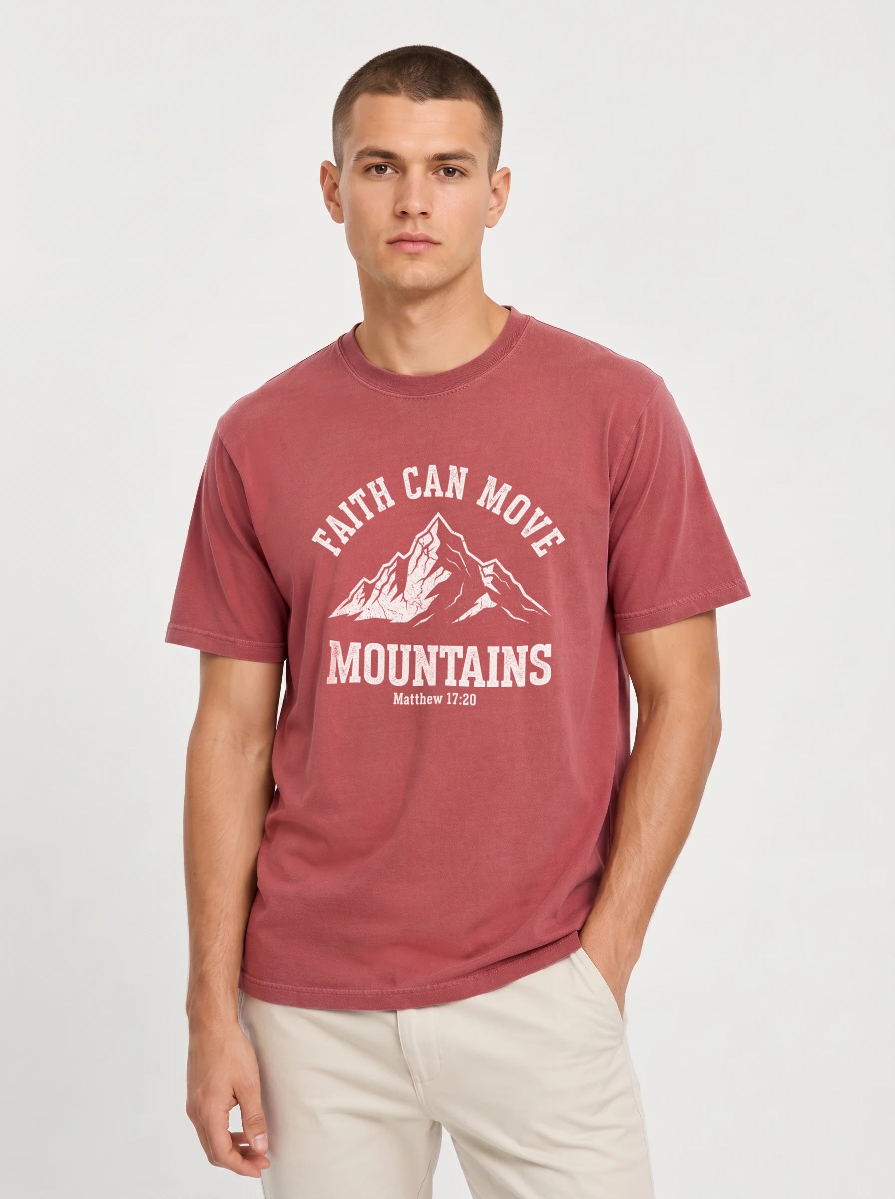 A man wearing a heathered dusty rose t-shirt with a white graphic print that reads 'FAITH CAN MOVE MOUNTAINS' and 'Matthew 17:20' below it, paired with light beige pants, against a light gray studio background.