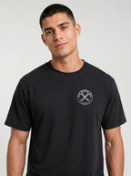 A man wearing a black crewneck t-shirt with a white circular graphic on the left side of the chest with the text 'IRON SHARPENS IRON' and 'PROVERBS 27:17' surrounding an illustration of two crossed nails, photographed against a light gray studio background.