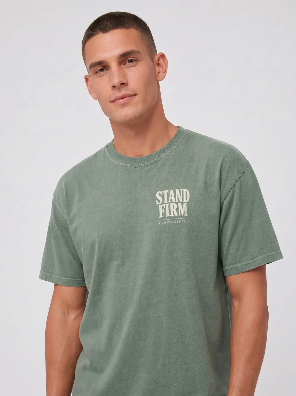 A man wearing a muted sage green crewneck t-shirt with a distressed cream-colored 'STAND FIRM' logo on the left chest, photographed against a neutral light gray background.