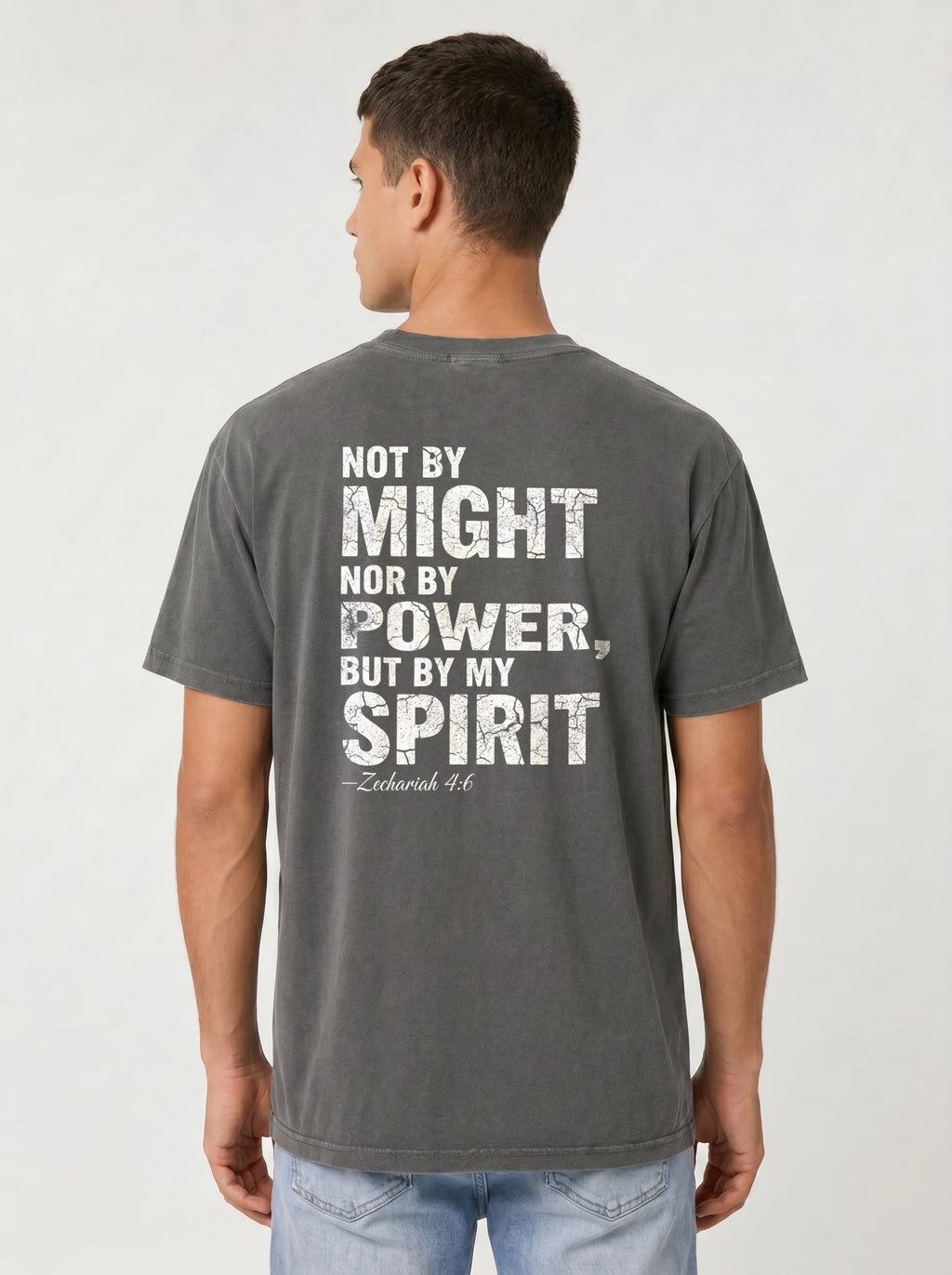 A man viewed from the back wearing a dark gray washed graphic t-shirt with a distressed white text print that reads 'NOT BY MIGHT NOR BY POWER, BUT BY MY SPIRIT -Zechariah 4:6'.