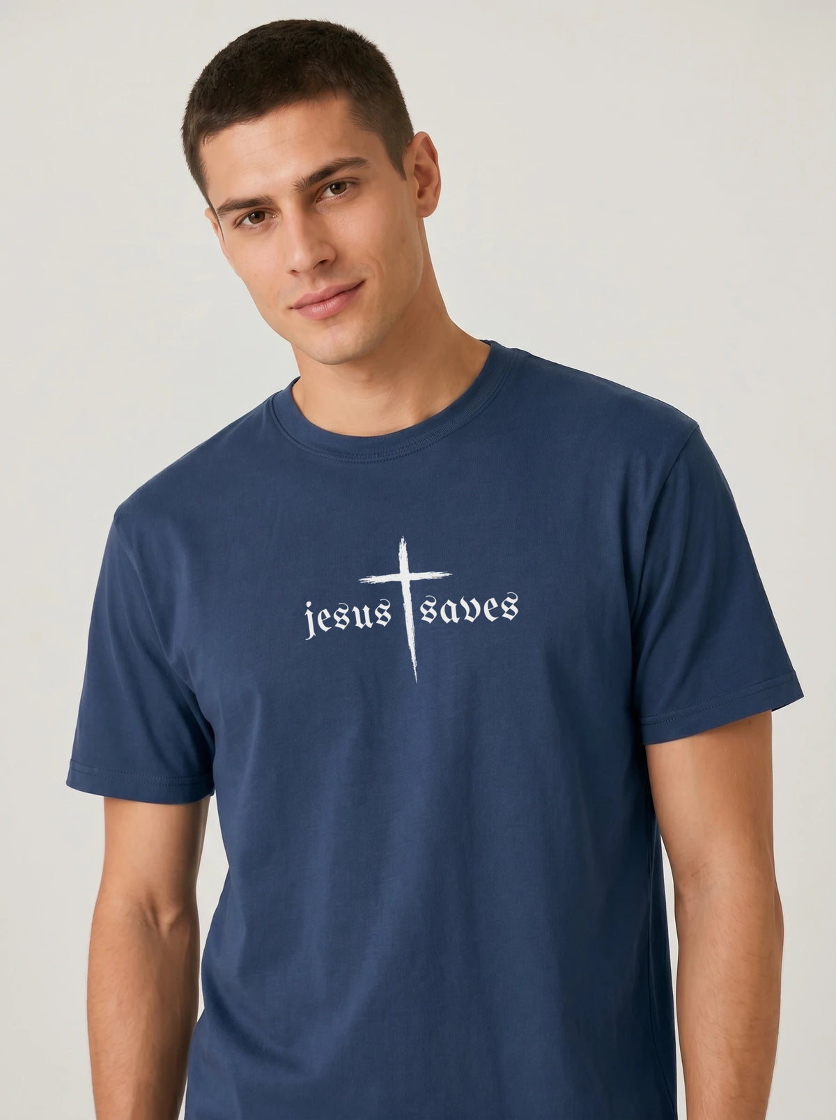 A man wearing a navy blue crewneck t-shirt with a white graphic on the chest that reads 'jesus saves' beneath a cross, set against a plain light gray background.