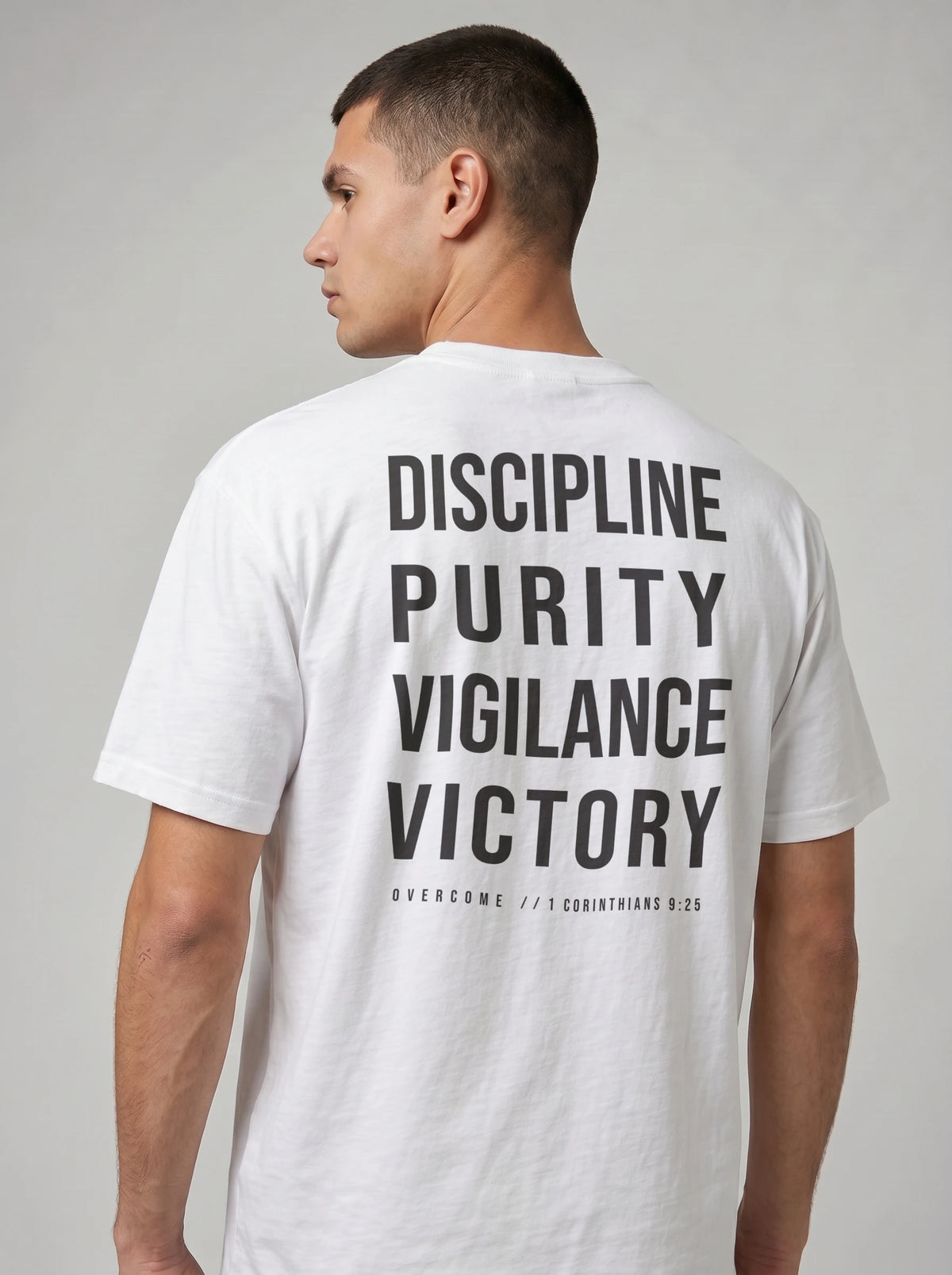 Person wearing a white T-shirt with large back text reading “Discipline Purity Vigilance Victory” and “Overcome // 1 Corinthians 9:25.”