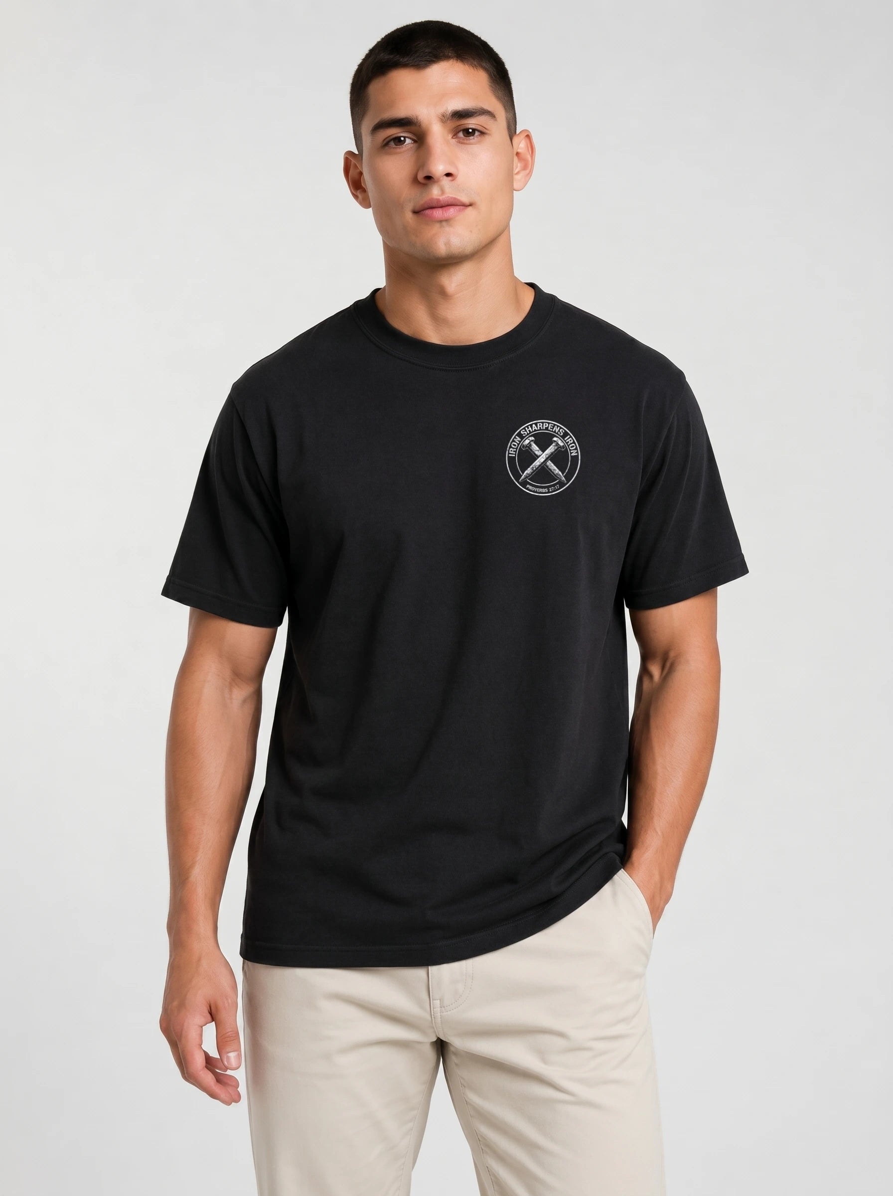 A man wearing a black short-sleeve crewneck t-shirt with a white circular graphic on the left side of the chest with the text 'IRON SHARPENS IRON' and 'PROVERBS 27:17' surrounding an illustration of two crossed nails, paired with beige pants, against a light gray studio background.