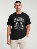 A man wearing a black short-sleeve t-shirt with a white graphic print featuring the quote 'NOT BY MIGHT NOR BY POWER, BUT BY MY SPIRIT' and a pair of boxing gloves, paired with beige pants, against a light gray background.