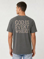 A man viewed from the back wearing a dark gray washed t-shirt with the text 'GOD IS EVERY WHERE' printed in a distressed beige font, paired with light blue jeans against a light gray studio background.