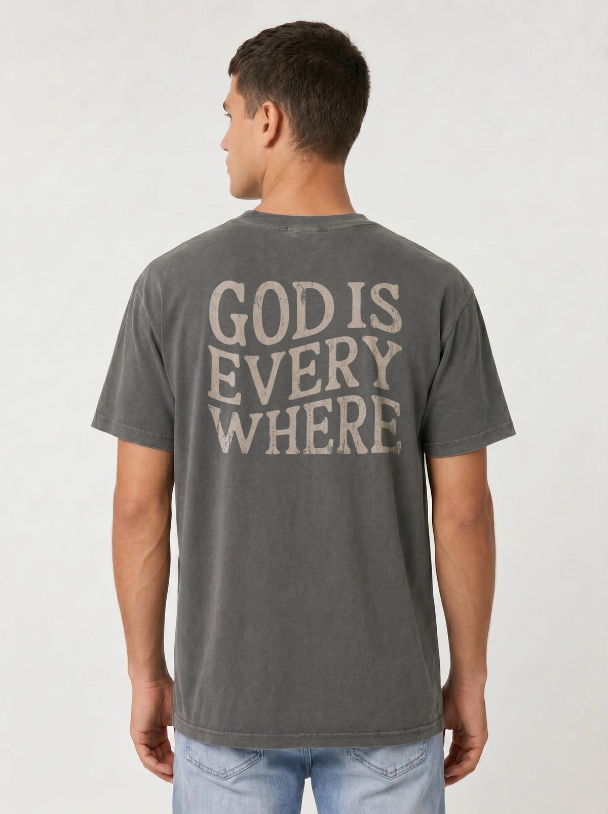 A man viewed from the back wearing a dark gray washed t-shirt with the text 'GOD IS EVERY WHERE' printed in a distressed beige font, paired with light blue jeans against a light gray studio background.