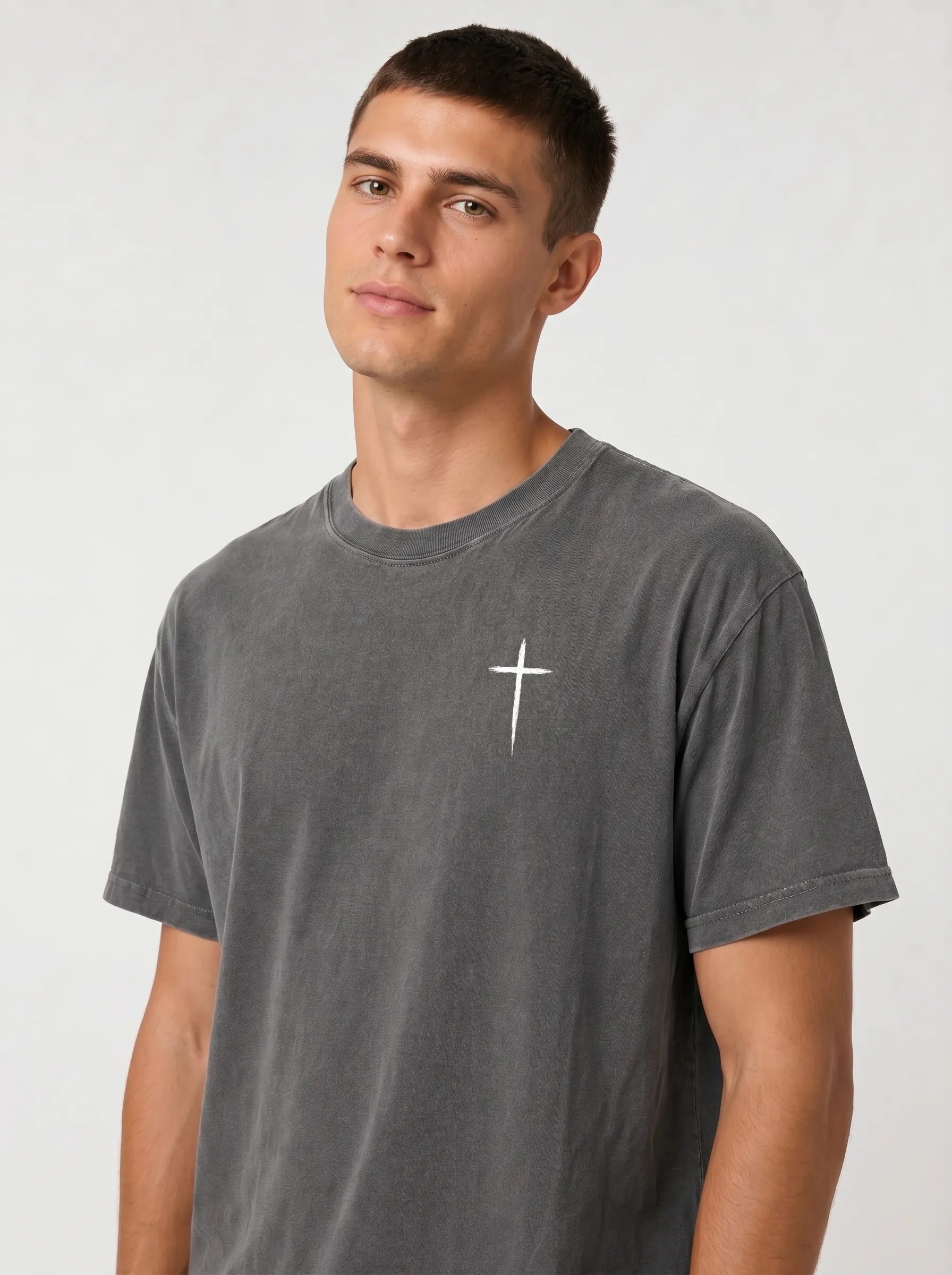 A man wearing a dark gray washed t-shirt with the text and a small cross printed on the chest, photographed against a light gray studio background.
