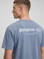 A man wearing a faded blue-gray t-shirt, viewed from the back. The shirt features the text 'Philippians 4:13' in a white Gothic font, with smaller white text underneath reading 'I CAN DO ALL THINGS THROUGH CHRIST WHO STRENGTHENS ME'.
