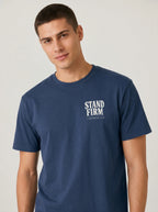 A man wearing a navy blue crewneck t-shirt with a white graphic print on the left chest that reads 'STAND FIRM'.