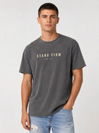 Person wearing a faded gray T-shirt with distressed beige text reading “Stand Firm” and “1 Cor. 16:13.”