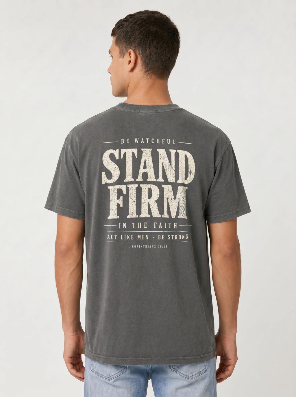 A man viewed from the back wearing a dark gray t-shirt with a distressed white graphic print that reads 'STAND FIRM IN THE FAITH' and light blue jeans, set against a plain light gray background.