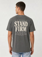 A man viewed from the back wearing a dark gray t-shirt with a distressed white graphic print that reads 'STAND FIRM IN THE FAITH' and light blue jeans, set against a plain light gray background.