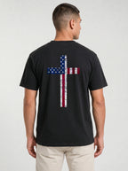 A man viewed from the back wearing a black t-shirt with a large distressed cross graphic in the American flag colors (red, white, and blue) on the back, paired with light beige pants, against a light gray studio background.