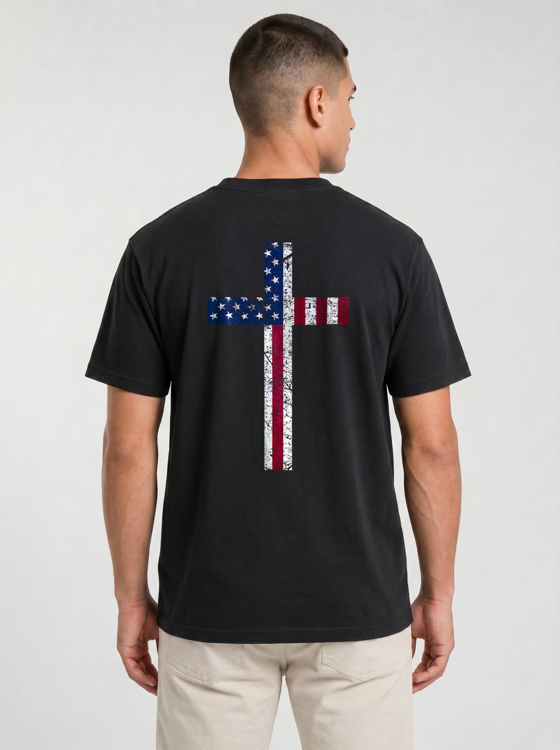 A man viewed from the back wearing a black t-shirt with a large distressed cross graphic in the American flag colors (red, white, and blue) on the back, paired with light beige pants, against a light gray studio background.