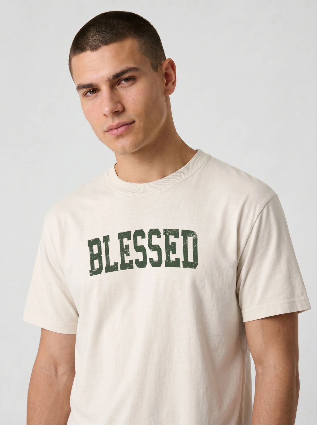 A man wearing a cream-colored t-shirt with the word 'BLESSED' printed across the chest in distressed dark green block letters, photographed against a light gray studio background.