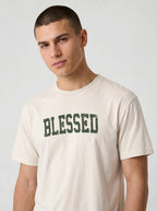 A man wearing a cream-colored t-shirt with the word 'BLESSED' printed across the chest in distressed dark green block letters, photographed against a light gray studio background.