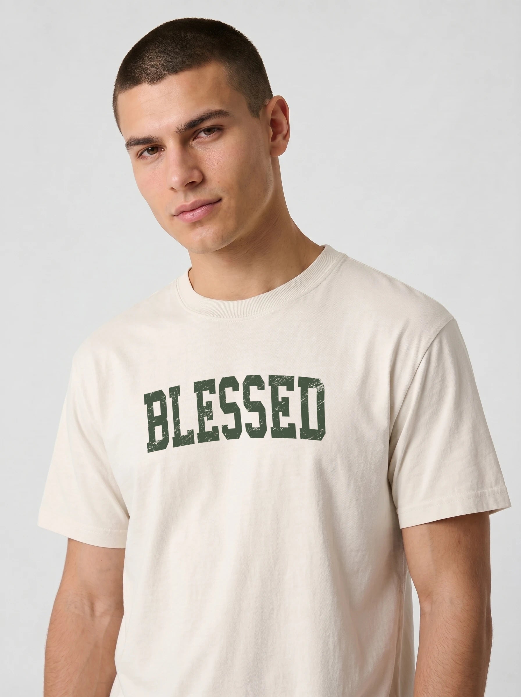 A man wearing a cream-colored t-shirt with the word 'BLESSED' printed across the chest in distressed dark green block letters, photographed against a light gray studio background.