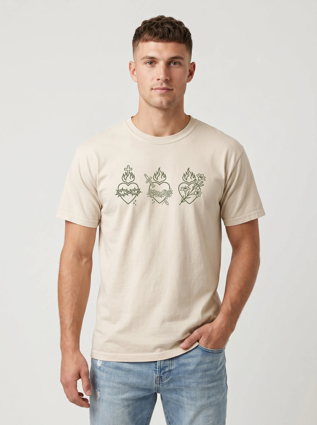 A man wearing a light beige crewneck t-shirt with a graphic of three sacred hearts in olive green, paired with light blue jeans, against a light gray studio background.