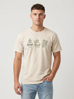 A man wearing a light beige crewneck t-shirt with a graphic of three sacred hearts in olive green, paired with light blue jeans, against a light gray studio background.