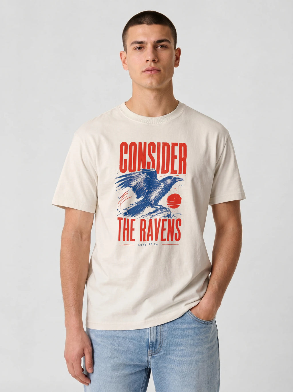 A man wearing a cream-colored graphic t-shirt with a red and blue raven design and the text 'CONSIDER THE RAVENS', paired with light blue jeans, against a light gray background.