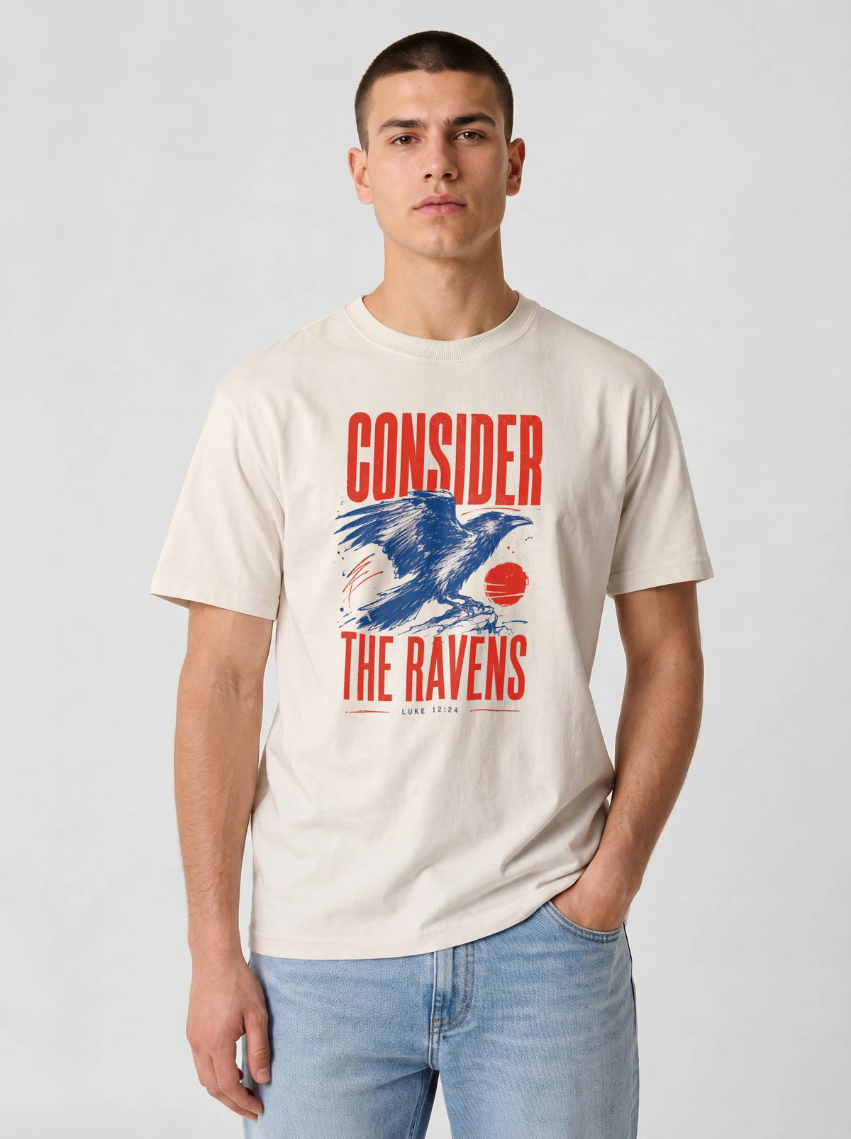 A man wearing a cream-colored graphic t-shirt with a red and blue raven design and the text 'CONSIDER THE RAVENS', paired with light blue jeans, against a light gray background.