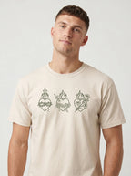A man wearing a light beige crewneck t-shirt with three Sacred Heart of Jesus line-art graphics printed across the chest, set against a plain light gray background.