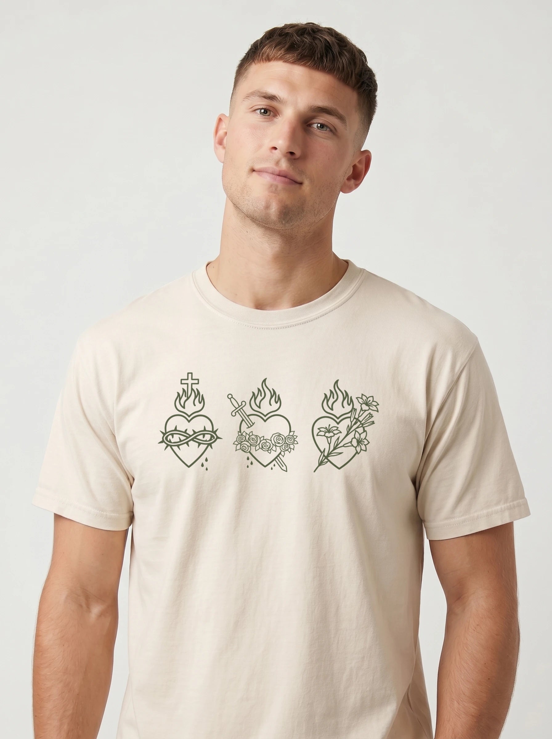 A man wearing a light beige crewneck t-shirt with three Sacred Heart of Jesus line-art graphics printed across the chest, set against a plain light gray background.
