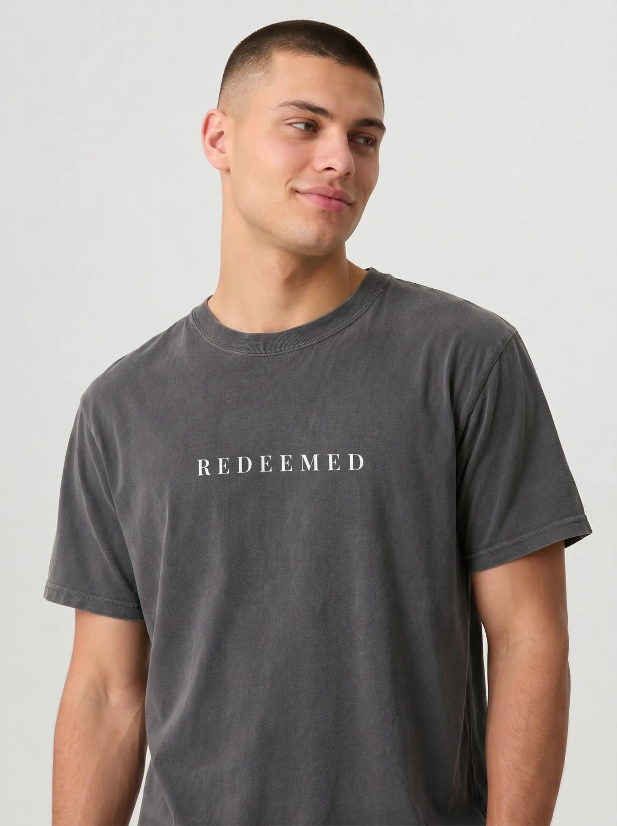 A man wearing a charcoal grey crewneck t-shirt with the word 'REDEEMED' printed in black serif letters across the chest, photographed against a light off-white background.