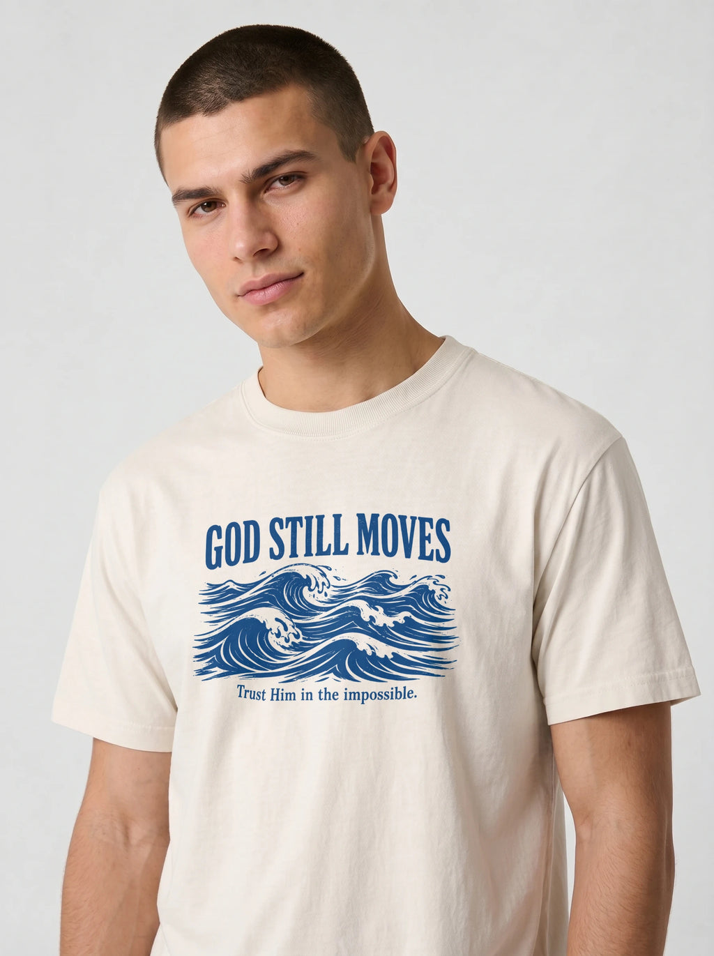 Close-up of a man's chest wearing a cream Comfort Colors t-shirt, showing the navy blue "GOD STILL MOVES" text above an ocean wave illustration and the phrase "Trust Him in the impossible."