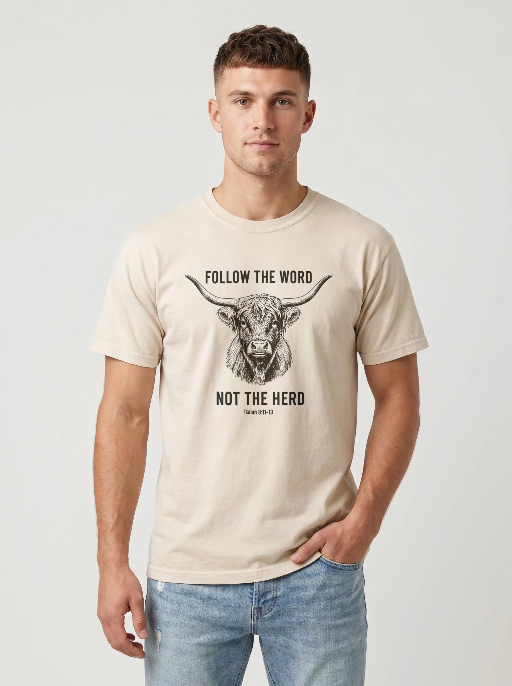 A man wearing a light beige crewneck t-shirt with a black graphic of a highland cow and the text 'FOLLOW THE WORD NOT THE HERD' and 'Isaiah 8:11-13', paired with light blue jeans, against a light gray studio background.