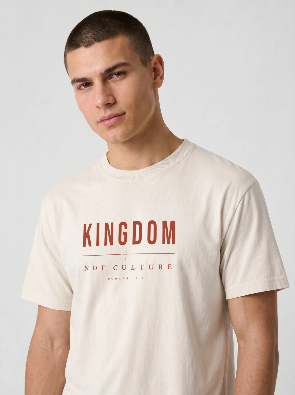 A man wearing a cream-colored short-sleeve t-shirt with a red graphic print that reads 'KINGDOM + NOT CULTURE' on the chest, set against a plain light gray background.