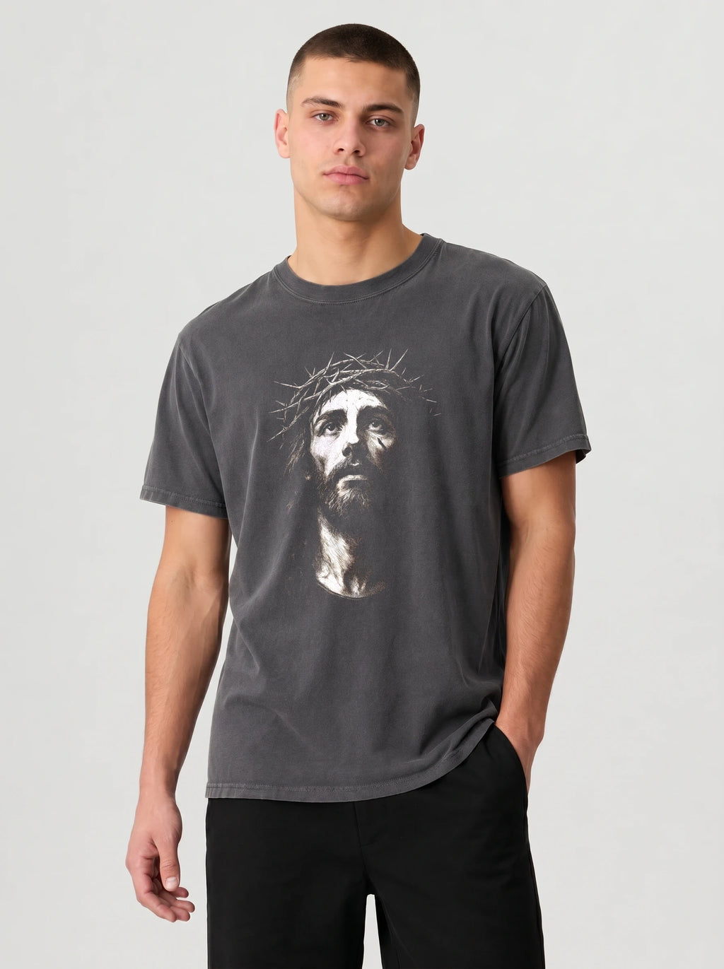 A man wearing a dark gray crewneck t-shirt with a white graphic print of Jesus' face on the front, paired with black pants, against a light gray studio background.
