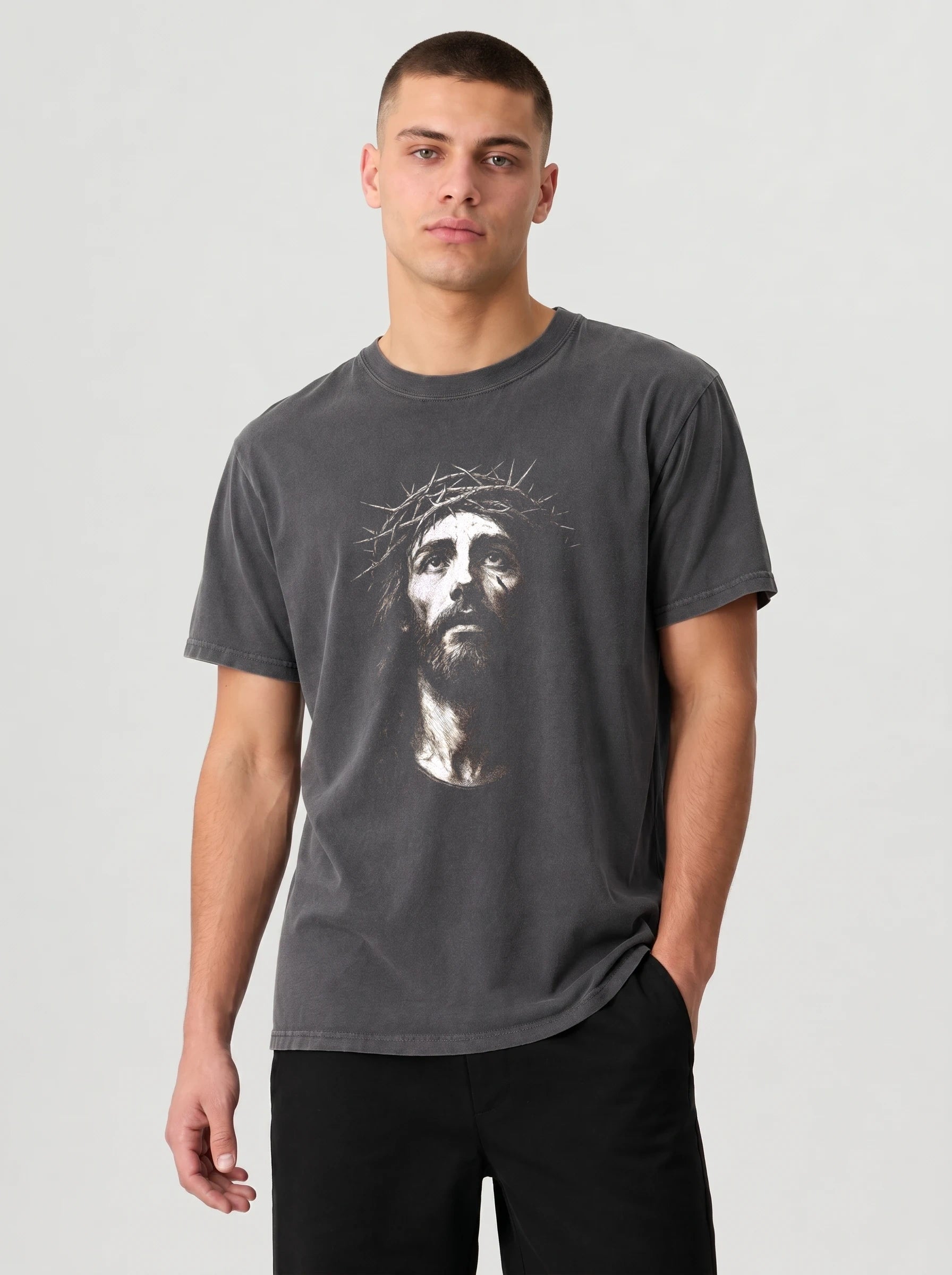 A man wearing a dark gray crewneck t-shirt with a white graphic print of Jesus' face on the front, paired with black pants, against a light gray studio background.