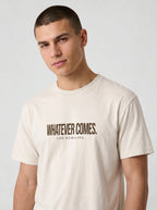 A man wearing a cream-colored t-shirt with the text 'WHATEVER COMES. GOD REMAINS.