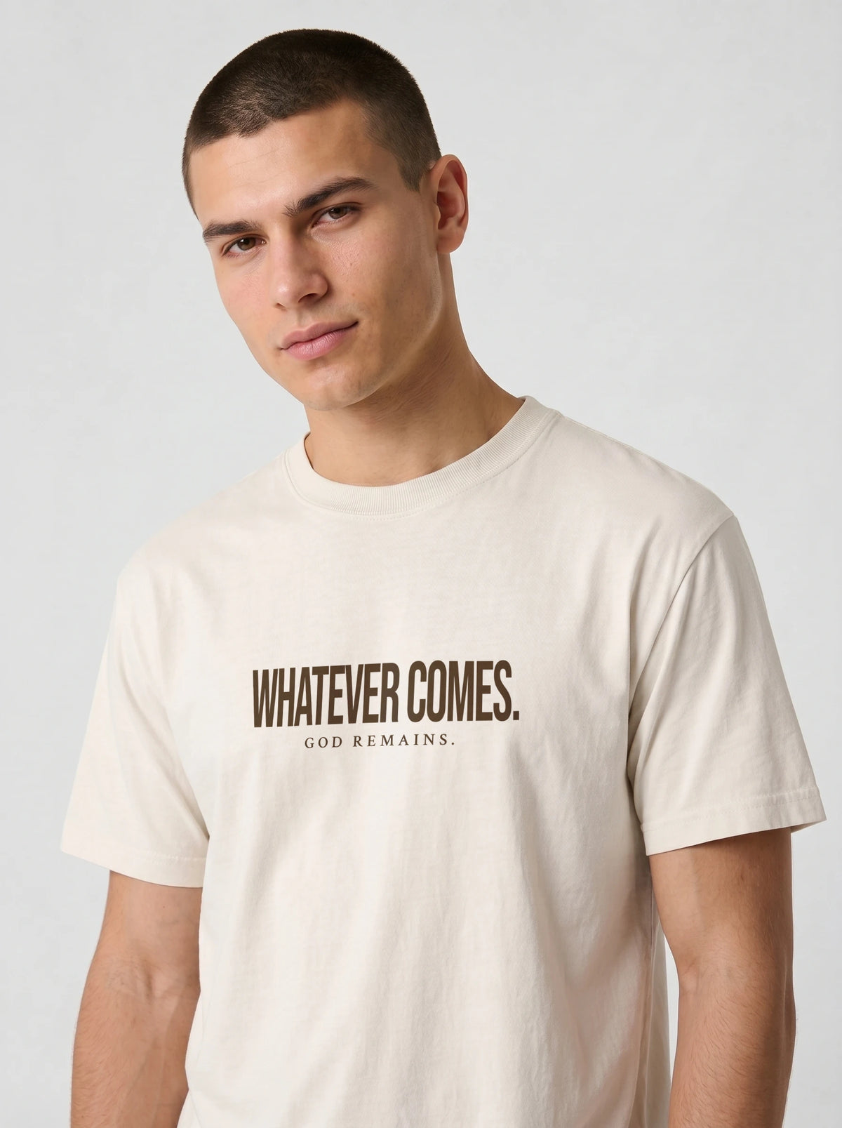 A man wearing a cream-colored t-shirt with the text 'WHATEVER COMES. GOD REMAINS.