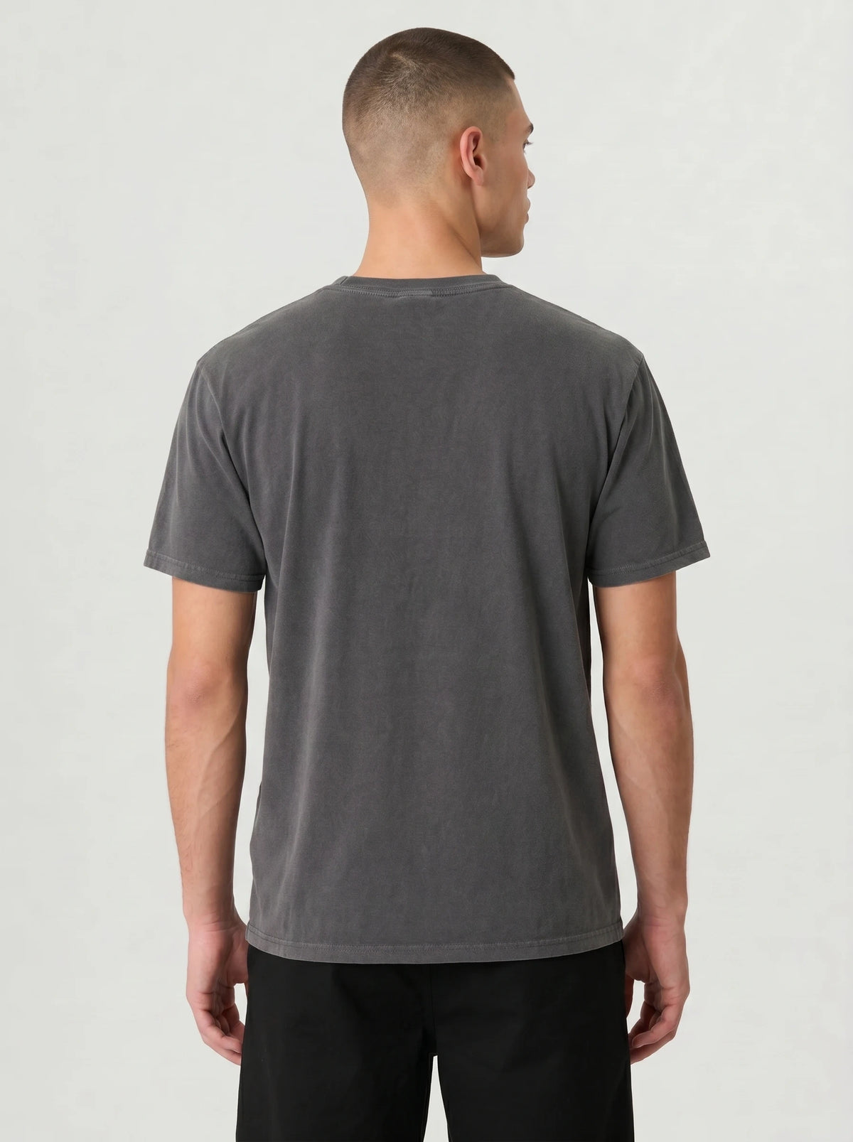 A man viewed from the back wearing a dark gray washed short-sleeve t-shirt and black pants, standing against a light gray studio background.