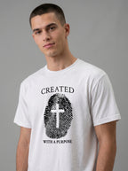 A man wearing a white crewneck t-shirt with a black graphic print that reads 'CREATED WITH A PURPOSE' surrounding a fingerprint design with a cross in the center, set against a plain gray studio background.