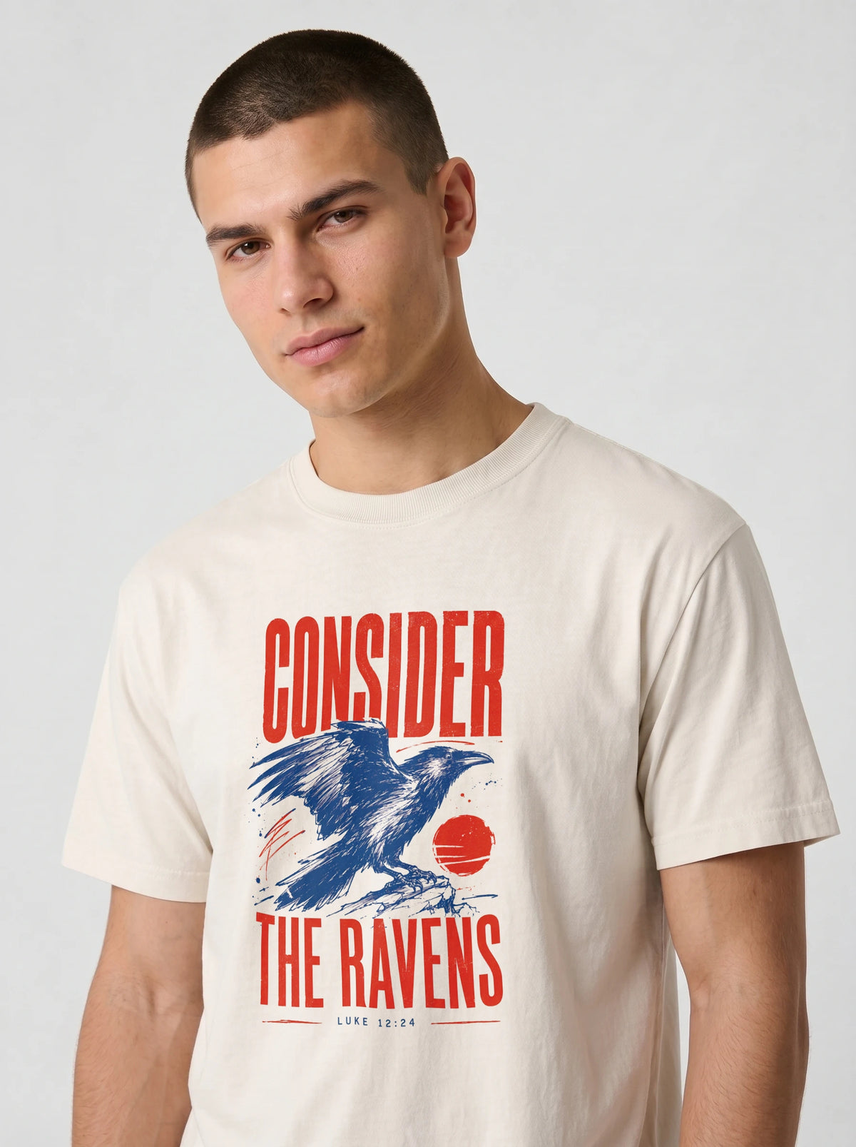 A man wearing a cream-colored graphic t-shirt with a red and blue raven design and the text 'CONSIDER THE RAVENS'.