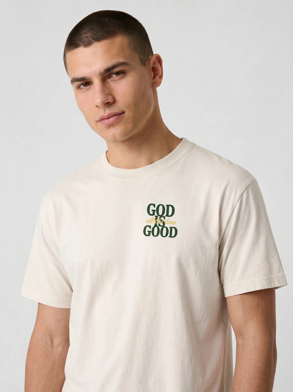 A man wearing a light beige crewneck t-shirt with a green graphic print on the chest that reads 'GOD IS GOOD'.