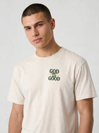 A man wearing a light beige crewneck t-shirt with a green graphic print on the chest that reads 'GOD IS GOOD'.