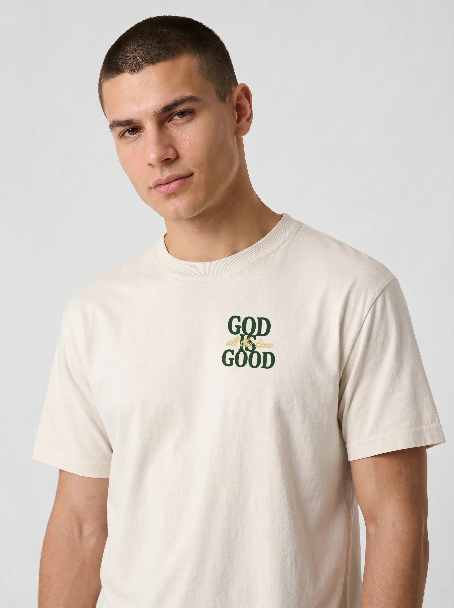 A man wearing a light beige crewneck t-shirt with a green graphic print on the chest that reads 'GOD IS GOOD'.
