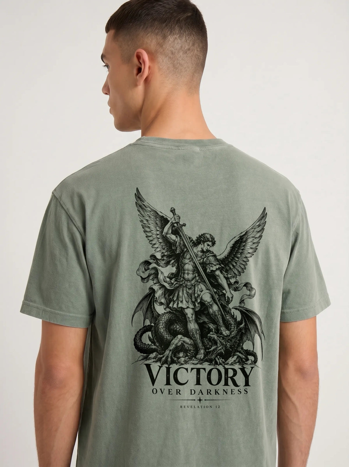 Sage green T-shirt with a winged warrior defeating a dragon and the text “VICTORY OVER DARKNESS” and “REVELATION 12.”