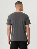 A man viewed from the back wearing a dark gray washed short-sleeve t-shirt and black pants, standing against a light gray studio background.