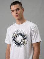 A man wearing a white crewneck t-shirt with a graphic print of a crown of thorns and white lilies, featuring Greek text.