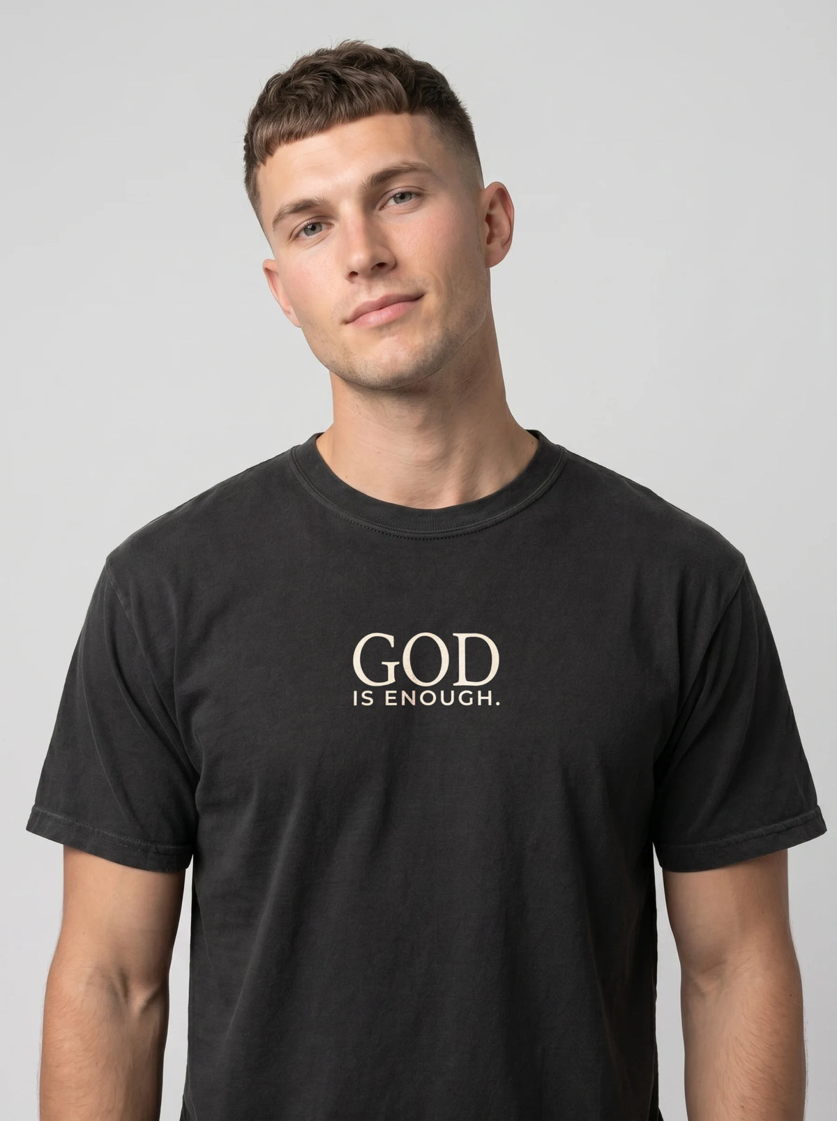 A man wearing a black crewneck t-shirt with the white printed text 'GOD IS ENOUGH. on the chest, photographed against a neutral gray background.