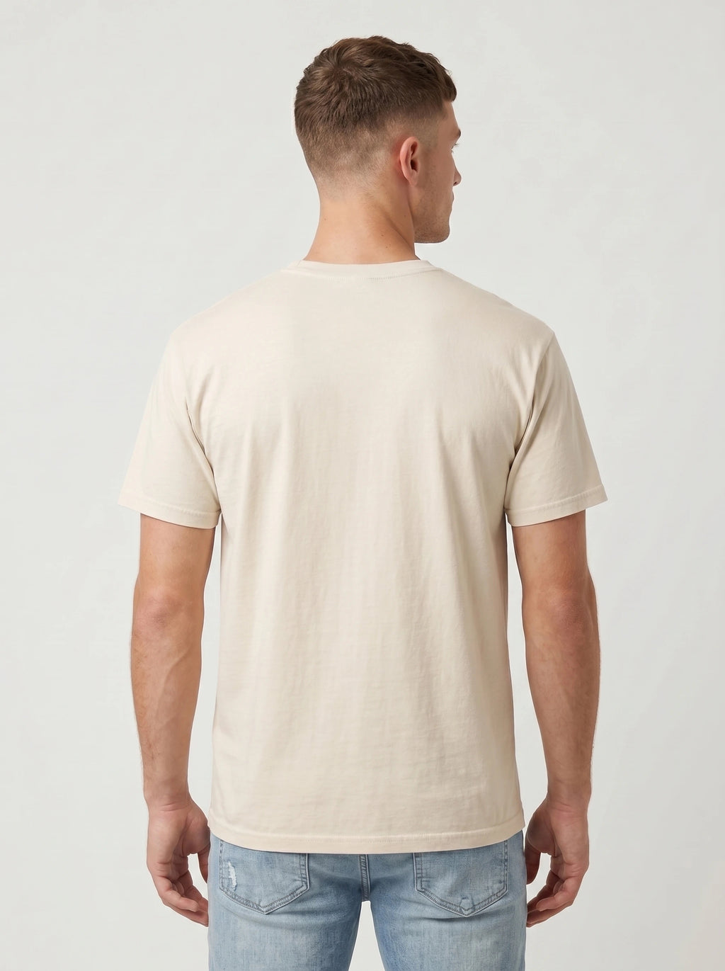 A man viewed from the back wearing a light beige crew-neck t-shirt and light blue jeans, standing against a plain light gray studio background.