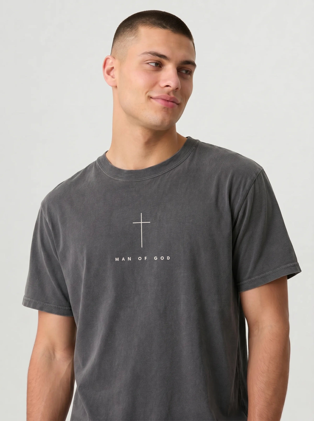A man wearing a dark gray washed t-shirt with a white cross and the text 'MAN OF GOD' printed on the chest, photographed against a light gray studio background.