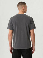 A man viewed from the back wearing a dark gray washed short-sleeve t-shirt and black pants, standing against a light gray studio background.