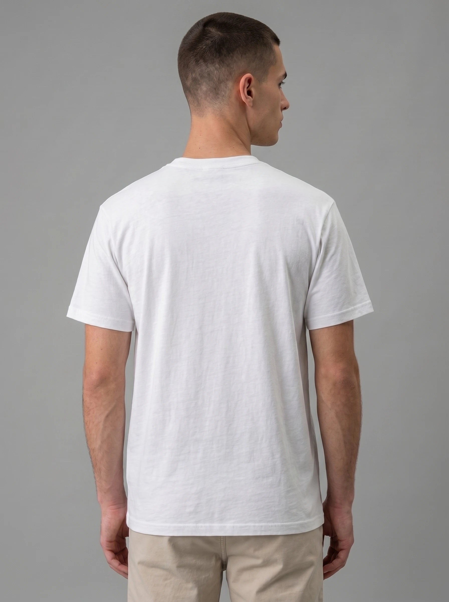 A man viewed from the back wearing a plain white short-sleeve crewneck t-shirt and beige pants, set against a solid gray studio background.